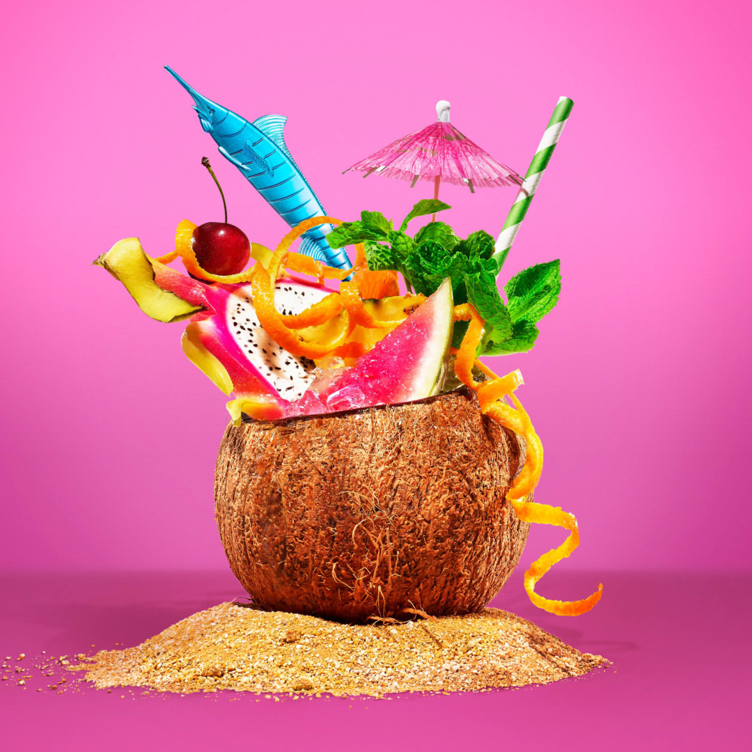 Tropical fruit cocktail in coconut shell with colorful accents, served on sand against a bright pink backdrop, vibrant and refreshing.