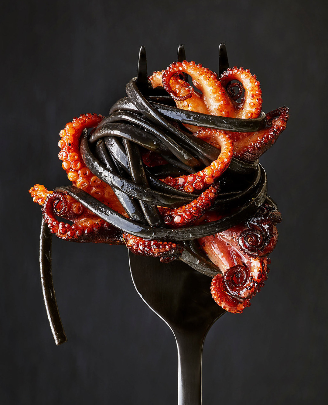 A fork holds a portion of black squid ink pasta and cooked octopus tentacles, presented against a dark background.