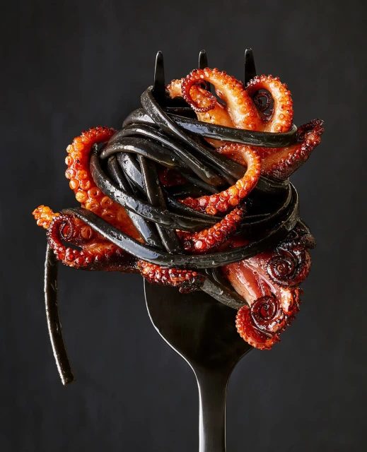 A fork holds a portion of black squid ink pasta and cooked octopus tentacles, presented against a dark background.