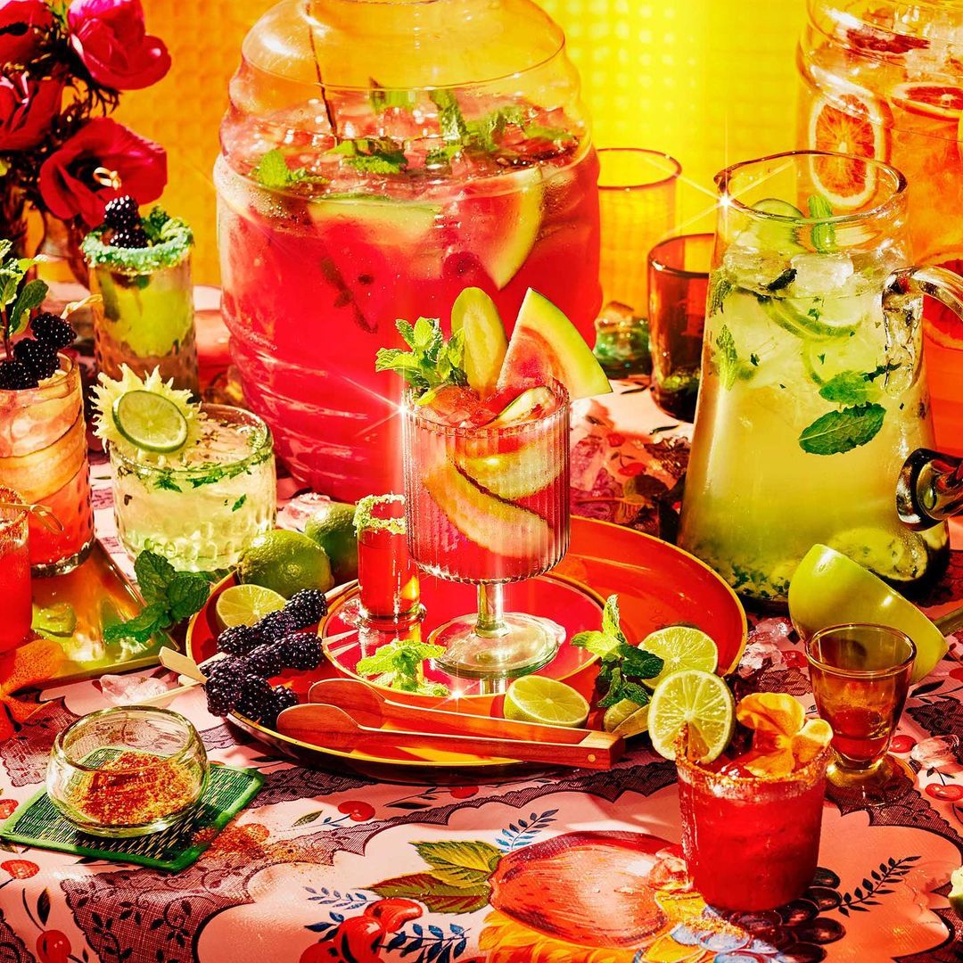Various cocktails and beverages arranged on a table with a floral tablecloth, featuring vibrant colors and fresh fruit garnishes.