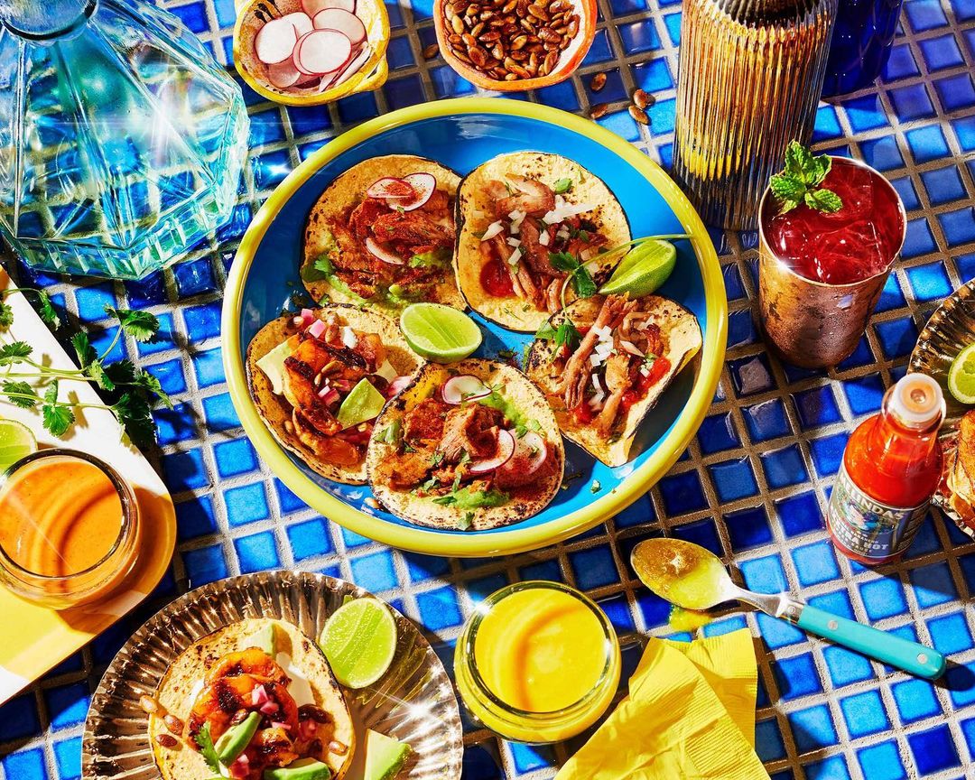 Overhead shot of assorted tacos on a blue-rimmed plate with limes, surrounded by various sauces and drinks on a blue-tiled surface.