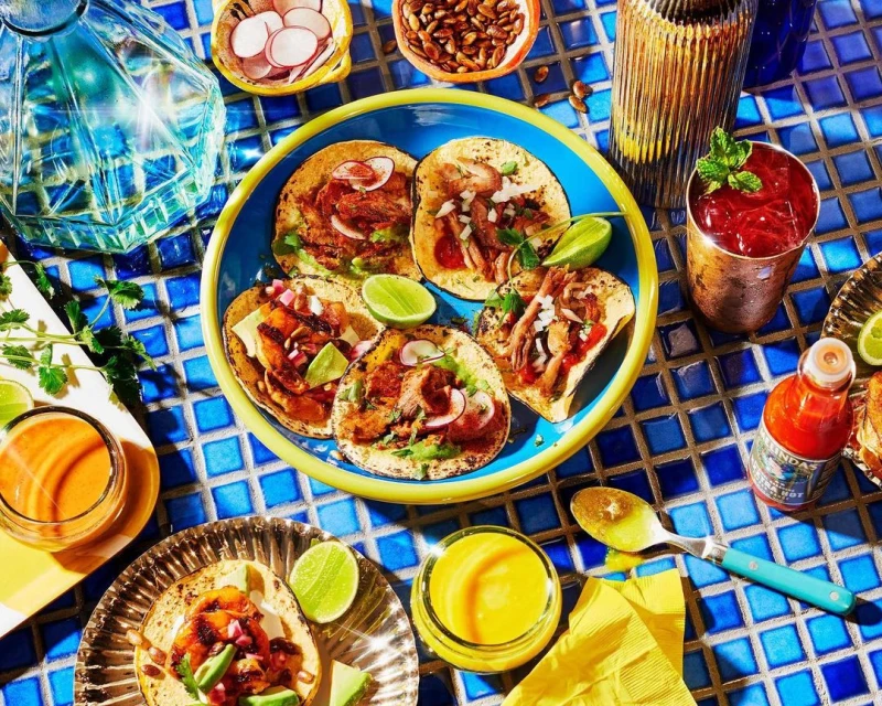 Overhead shot of assorted tacos on a blue-rimmed plate with limes, surrounded by various sauces and drinks on a blue-tiled surface.