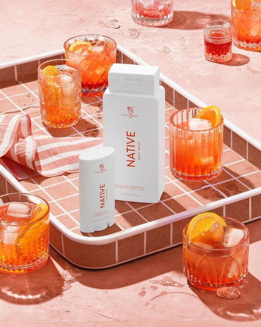 Two white bottles of Native brand body wash and deodorant are staged with multiple glasses of orange cocktails on a tile tray.