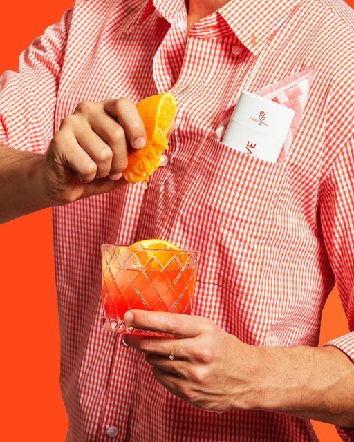 Person squeezing orange into glass with cocktail; drink in glass in hand; sleeve and handkerchief in shirt pocket.