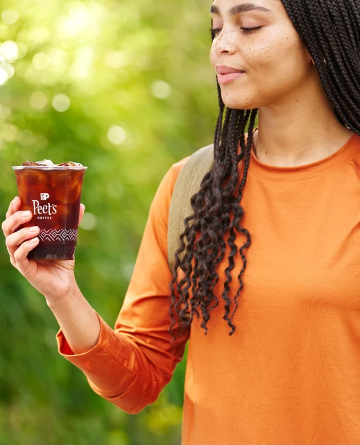 A woman in an orange shirt holds a Peet's iced coffee cup outside, with a blurred green background. The cup and woman are the focal point.