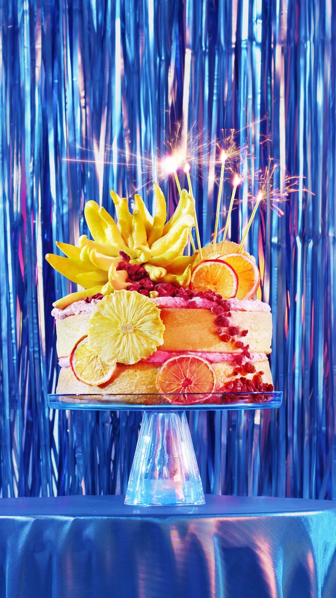 Celebratory cake with yellow flower and citrus fruit topping, set on a clear stand against a blue tinsel backdrop with sparklers.