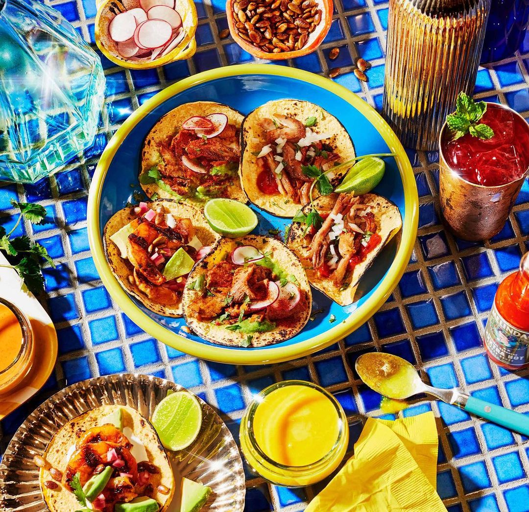 Overhead view of six tacos arranged on a bright blue plate, surrounded by garnishes, sauces, and a drink, set on a blue tile surface.