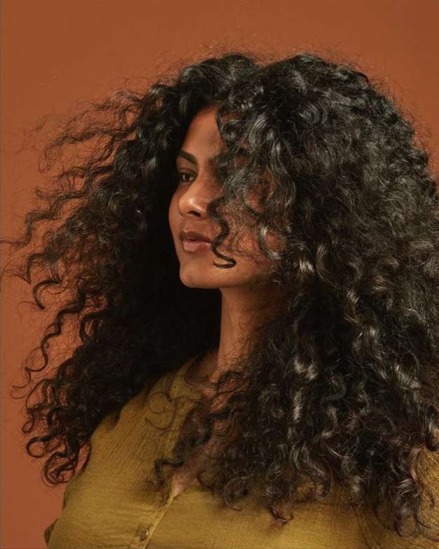 Portrait of an adult woman with long, voluminous, dark curly hair wearing a mustard-yellow top against a warm brown background.