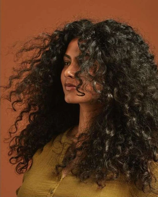 Portrait of an adult woman with long, voluminous, dark curly hair wearing a mustard-yellow top against a warm brown background.