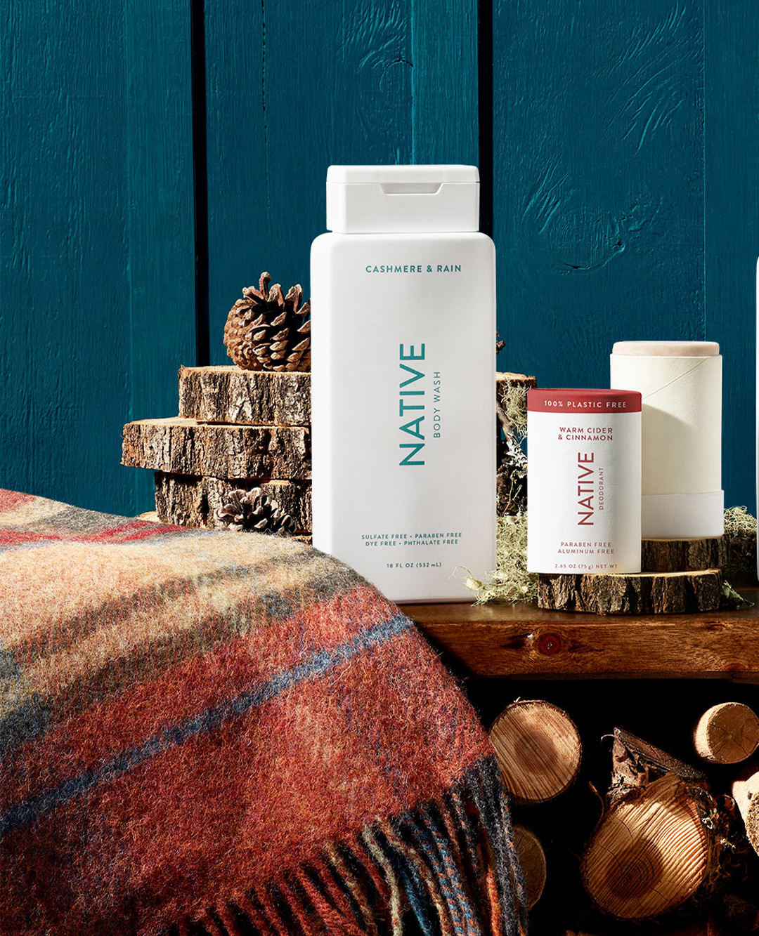 Native brand body wash, deodorant products displayed on wood with pinecone and patterned throw against a blue paneled background.