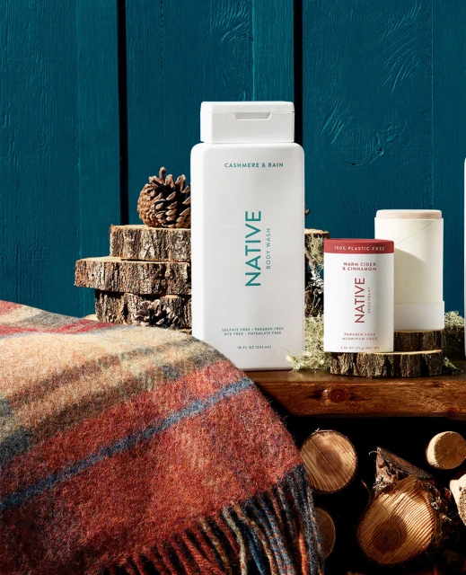 Native brand body wash, deodorant products displayed on wood with pinecone and patterned throw against a blue paneled background.