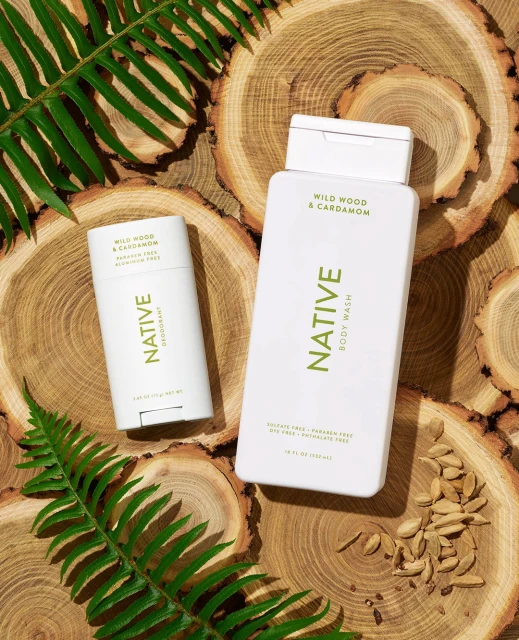 Product shot of Native brand body wash and deodorant on wood slices with green ferns, emphasizing natural, eco-friendly personal care.