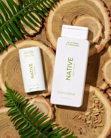 Product shot of Native brand body wash and deodorant on wood slices with green ferns, emphasizing natural, eco-friendly personal care.