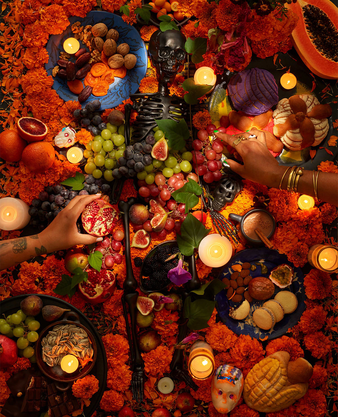 Overhead shot of a Day of the Dead altar with marigolds, skull, candles, fruit, bread, and hands adding items to the display.