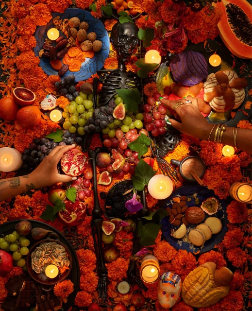 Overhead shot of a Day of the Dead altar with marigolds, skull, candles, fruit, bread, and hands adding items to the display.