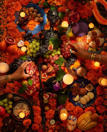 Overhead shot of a Day of the Dead altar with marigolds, skull, candles, fruit, bread, and hands adding items to the display.