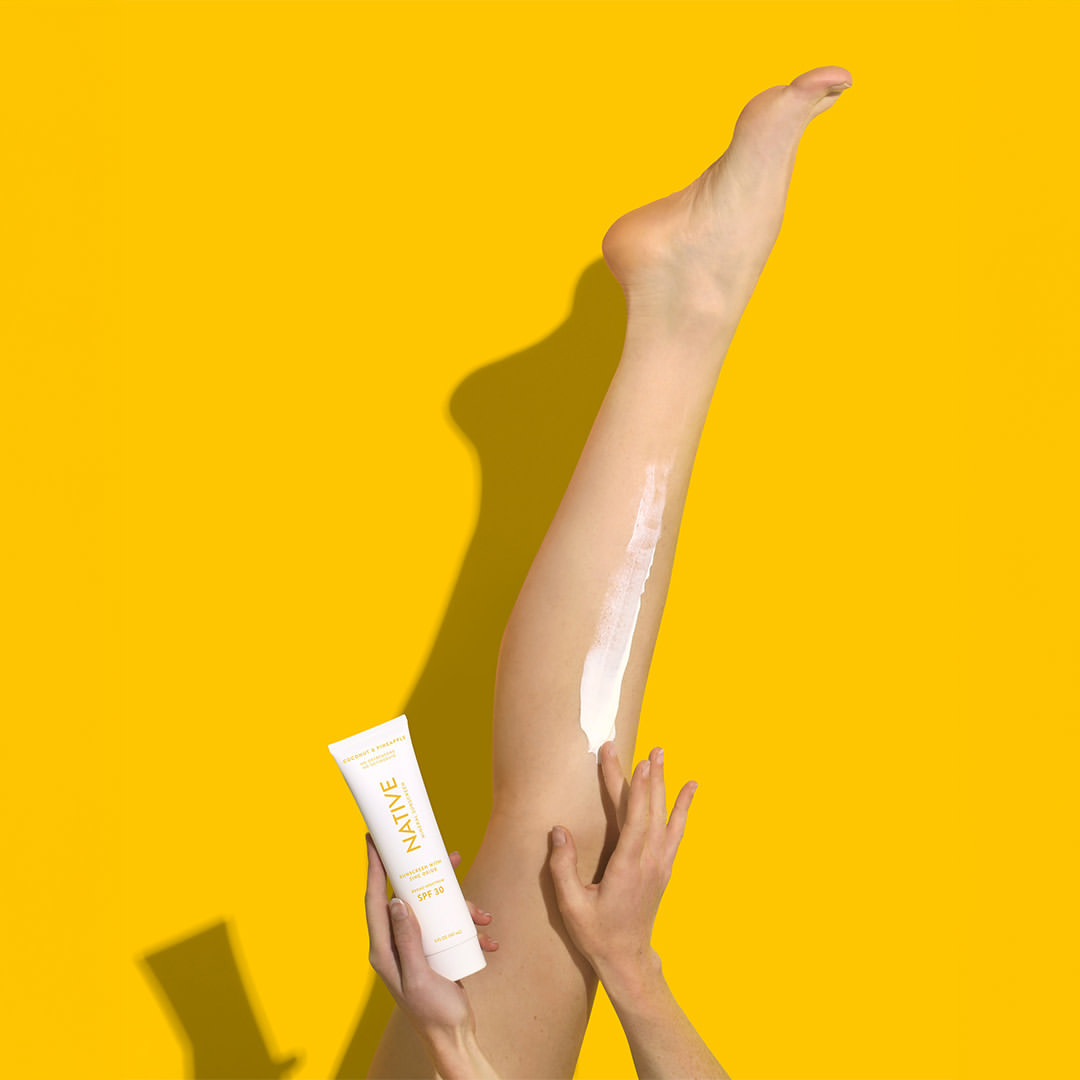 Close-up of a woman applying cream to her leg with a tube of Native sunscreen against a bright yellow studio backdrop.