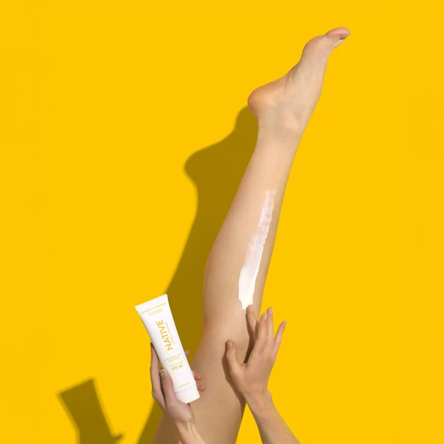 Close-up of a woman applying cream to her leg with a tube of Native sunscreen against a bright yellow studio backdrop.