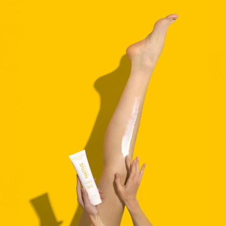 Close-up of a woman applying cream to her leg with a tube of Native sunscreen against a bright yellow studio backdrop.