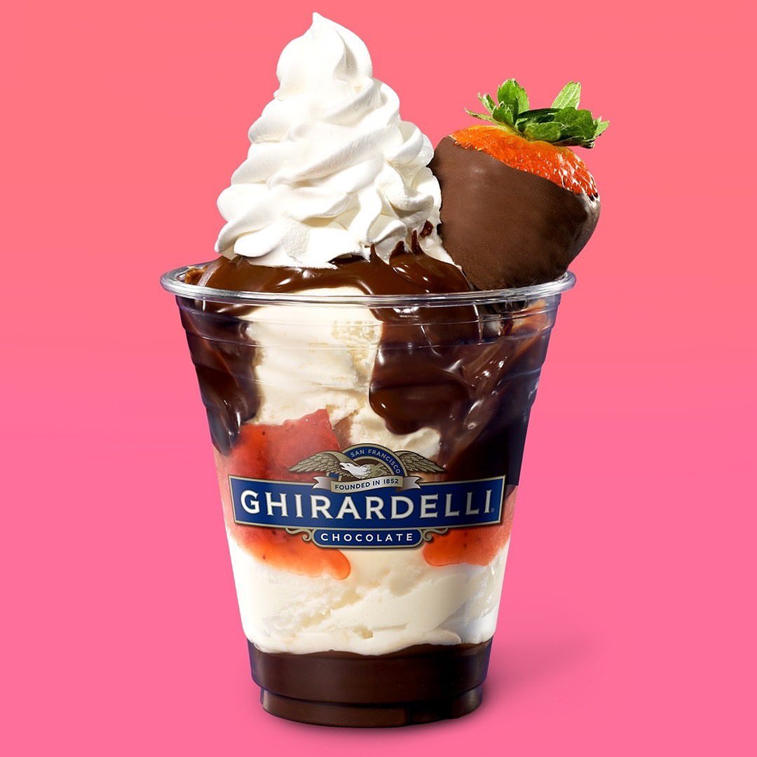 Ghirardelli ice cream sundae with whipped cream, chocolate sauce, and a chocolate-covered strawberry against a pink background.