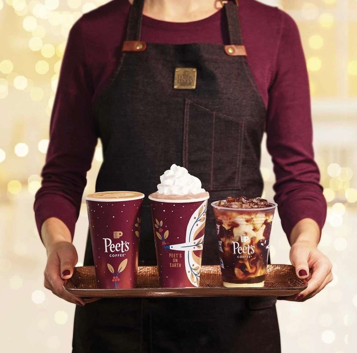 A person in an apron holds a tray with three Peet's Coffee beverages, one with whipped cream, against a bokeh background of holiday lights.