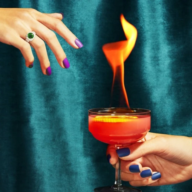 A cocktail glass with flaming beverage, held by a manicured hand, and another hand with jewelry. Dramatic lighting on teal background.