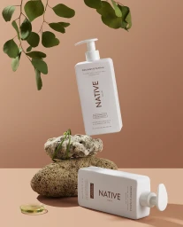 Two white Native shampoo and conditioner bottles stacked with rocks and eucalyptus leaves on a warm brown background