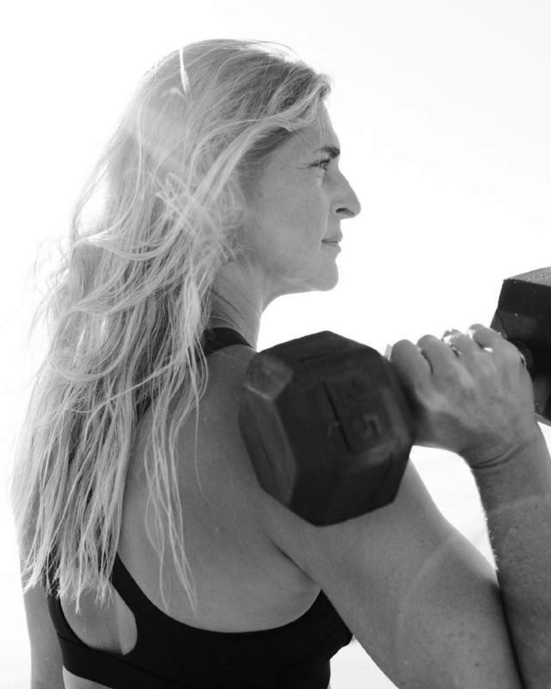 Black and white portrait of an adult female in sportswear lifting a dumbbell with her hair flowing, fitness and exercise theme.