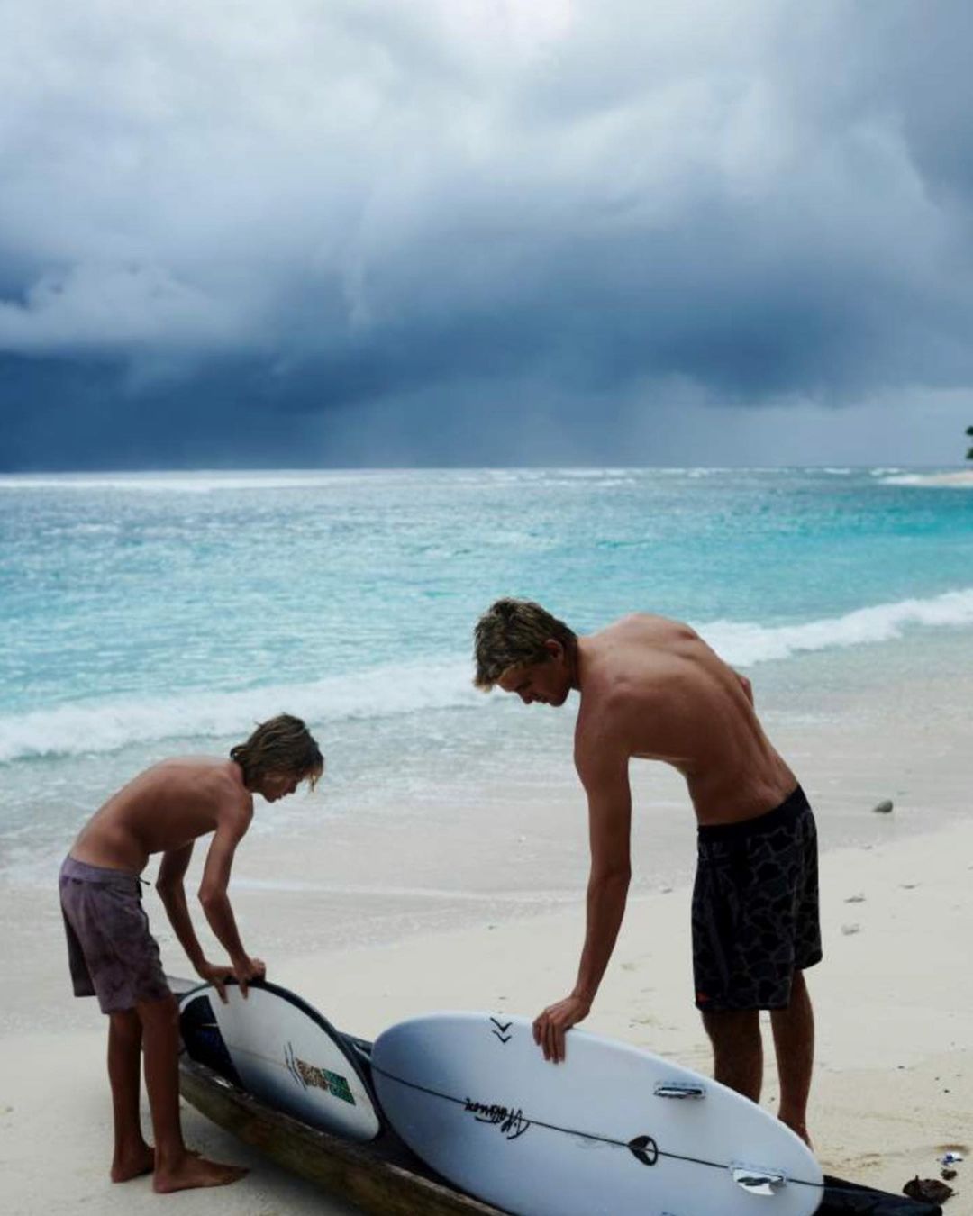 Two surfers with boards on a beach under a cloudy sky, preparing for a surf session on the turquoise ocean water.