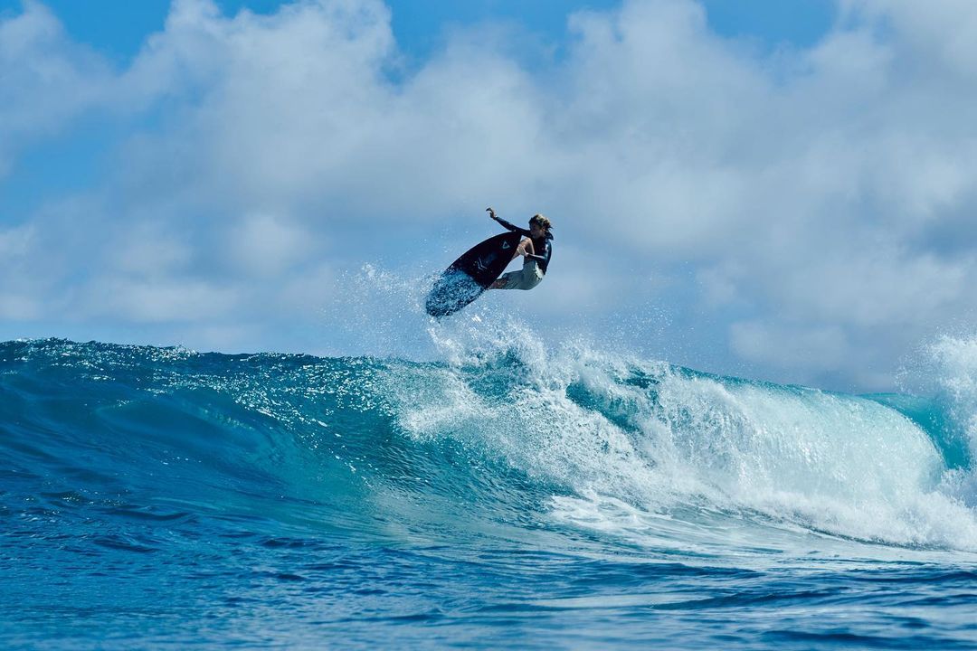 A surfer catches air on a blue wave against a partly cloudy sky, showcasing skill in an ocean setting.