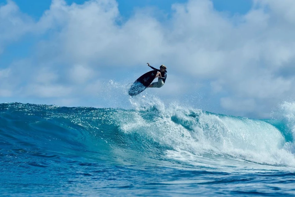 A surfer catches air on a blue wave against a partly cloudy sky, showcasing skill in an ocean setting.