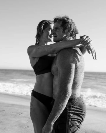 Black and white portrait of a couple embracing on a sandy beach with the ocean in the background by photographer Anne Menke.