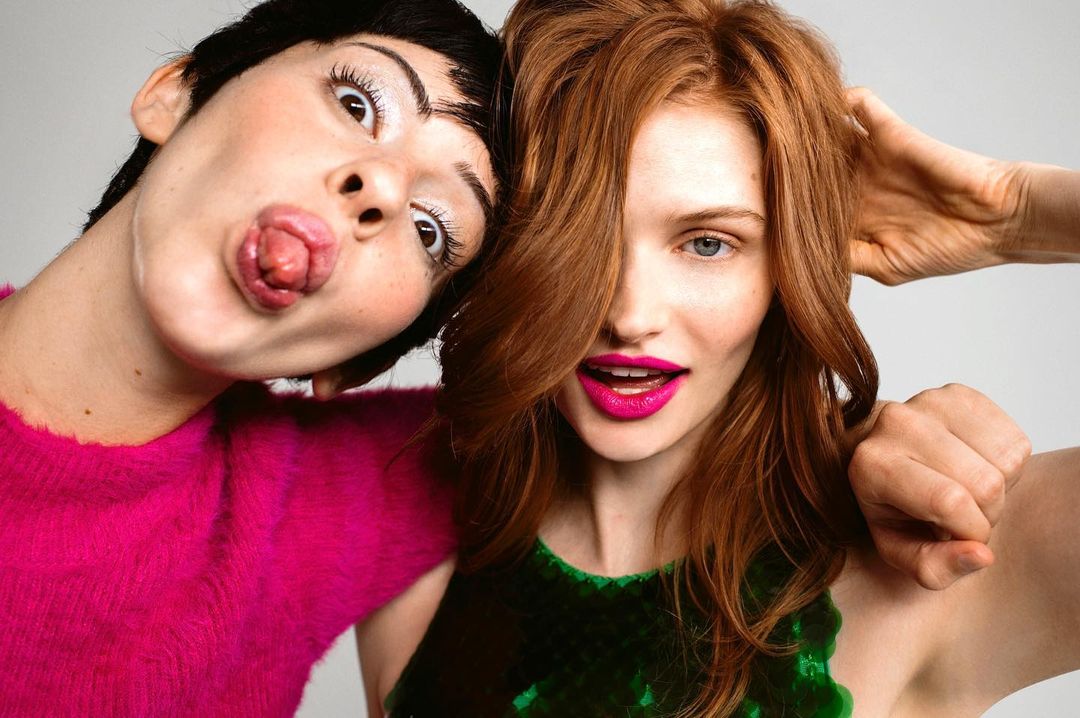 Two playful women pose closely in a studio shot. One has short dark hair and sticks her tongue out. The other, a redhead, smiles brightly.