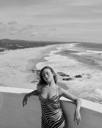 Black and white photo of a woman in a zebra-striped dress leaning against a wall, with the ocean and beach in the background.