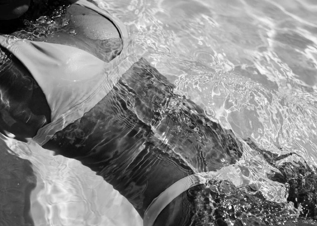 Monochrome close-up shows a woman wearing a bikini in water with refracted light patterns. Dominant colors are shades of black, white, and gray.
