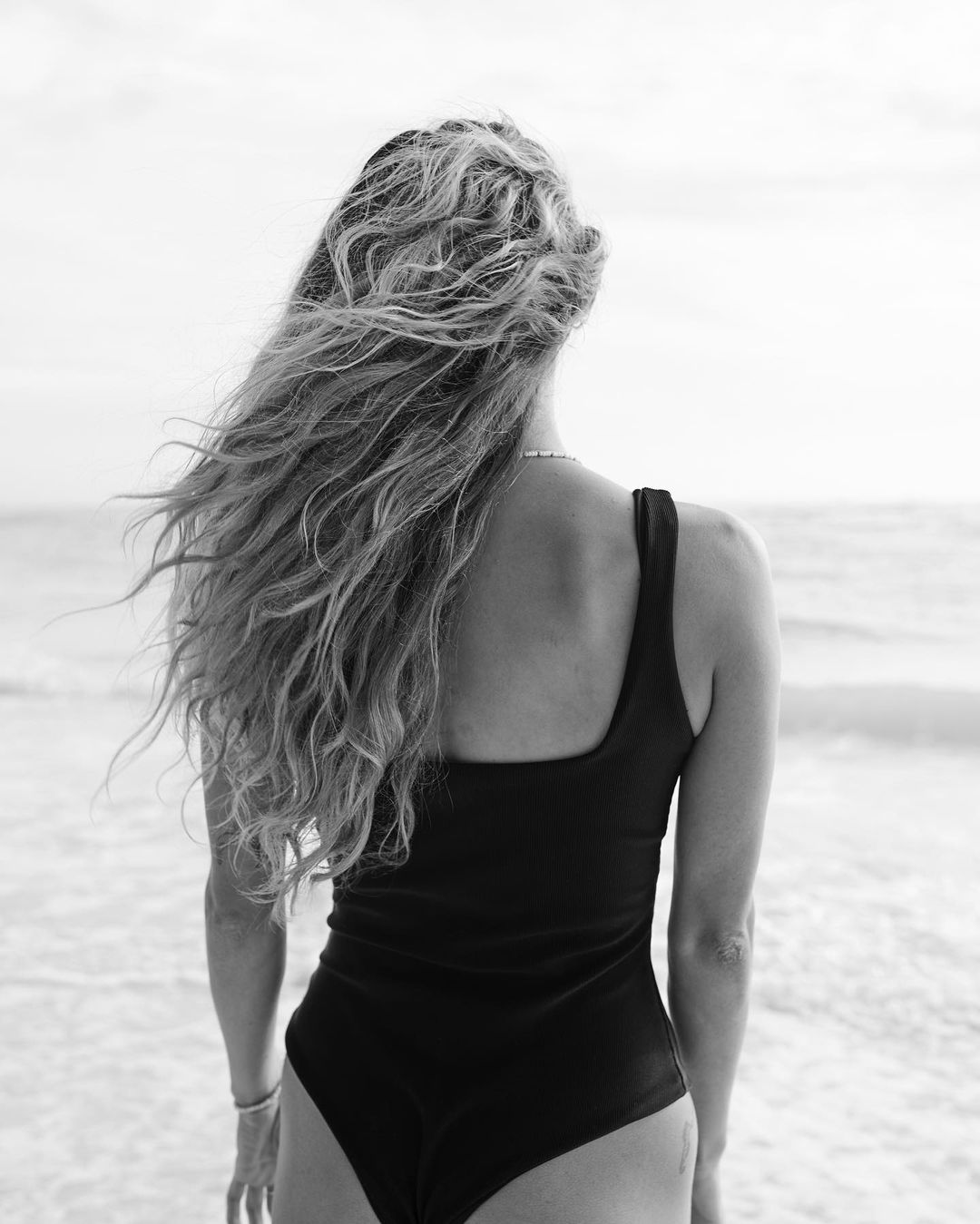 Woman with long curly hair seen from behind wearing a dark one-piece swimsuit on a sandy beach by the ocean.