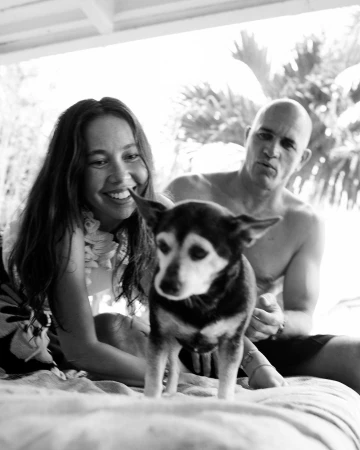 Black and white photo of a smiling woman, a dog, and a man, all sitting outdoors with greenery visible in the background on a sunny day.