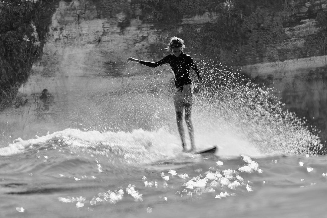 Black and white action shot of a surfer riding a wave on a surfboard, showcasing water splashing and a cliffside in the background.