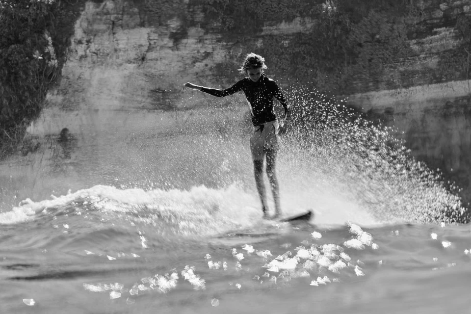 Black and white action shot of a surfer riding a wave on a surfboard, showcasing water splashing and a cliffside in the background.