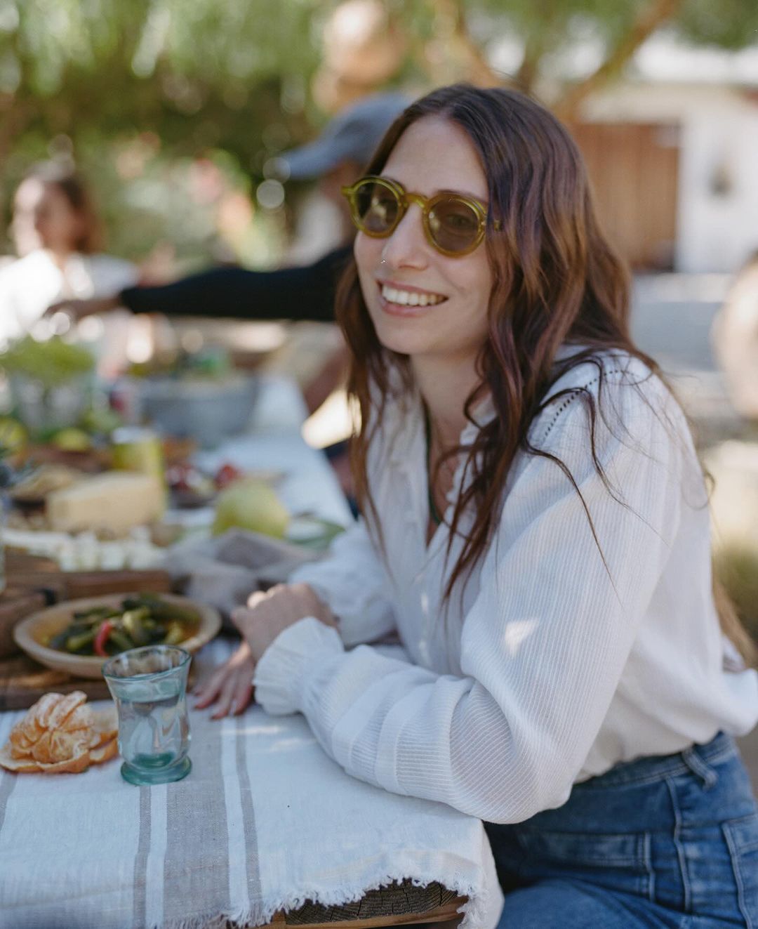 A woman with long brown hair smiles at an outdoor table covered in food and drinks, wearing olive sunglasses and a white blouse.