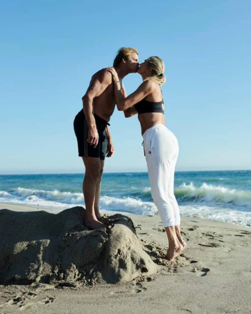 Mature couple kissing on sandy beach with ocean waves and blue sky. Barefoot man and woman wearing athletic clothing share affection outdoors.