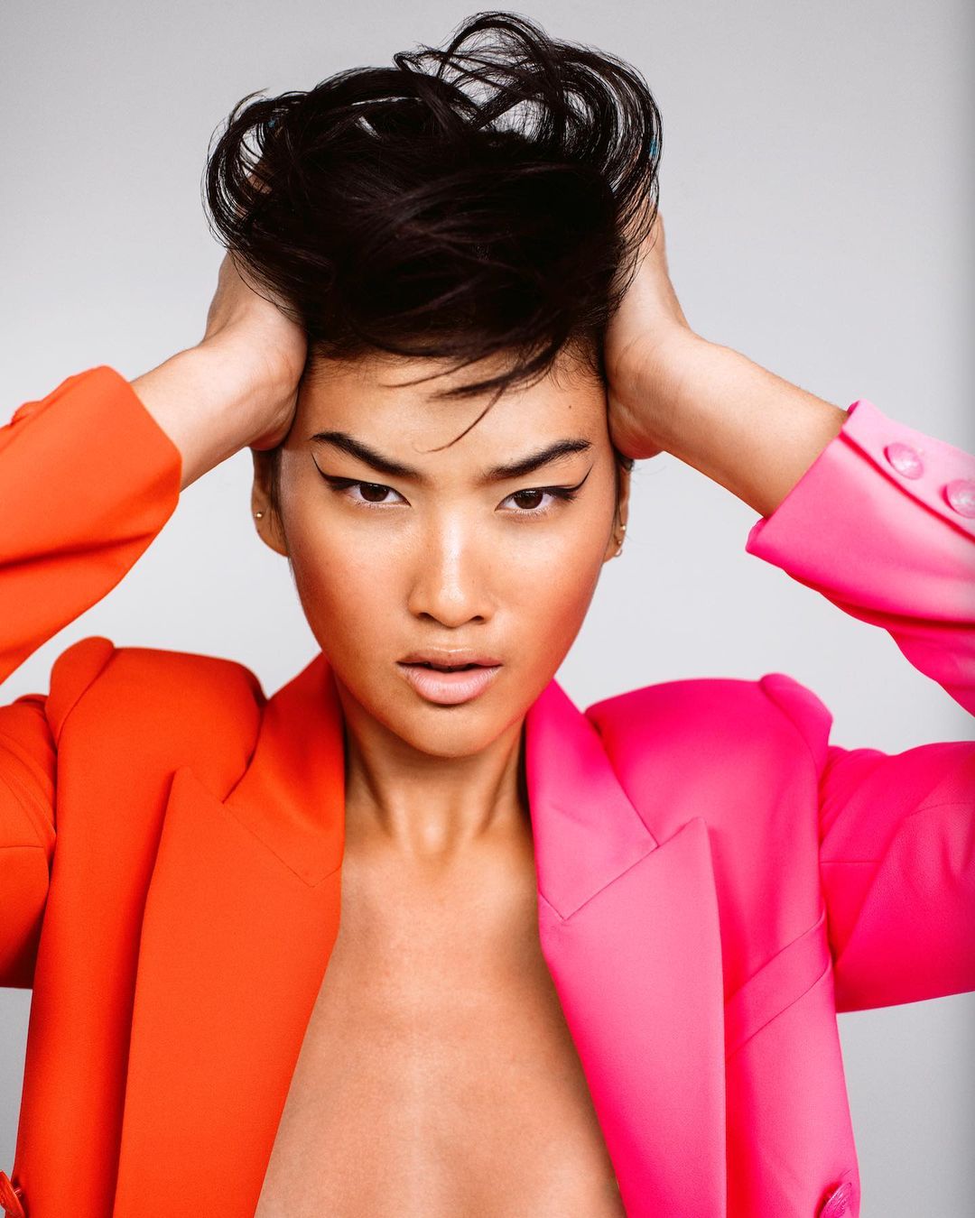 Close-up portrait of a woman with dark hair holding her head, wearing an orange and pink blazer against a plain light background.