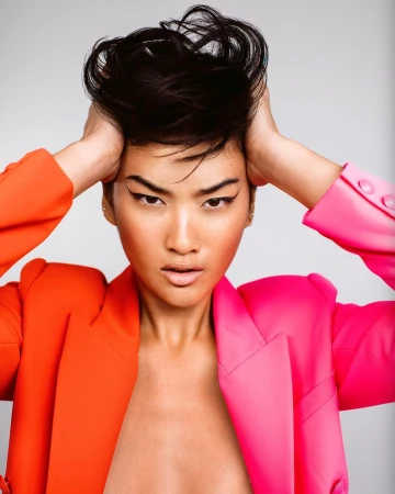 Close-up portrait of a woman with dark hair holding her head, wearing an orange and pink blazer against a plain light background.