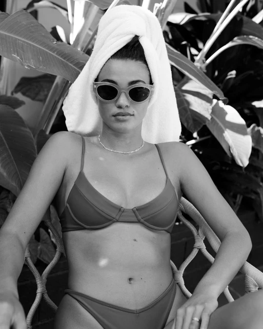 A young woman wearing sunglasses and a bikini with a towel wrapped around her head, poses seated in a chair with tropical plants.