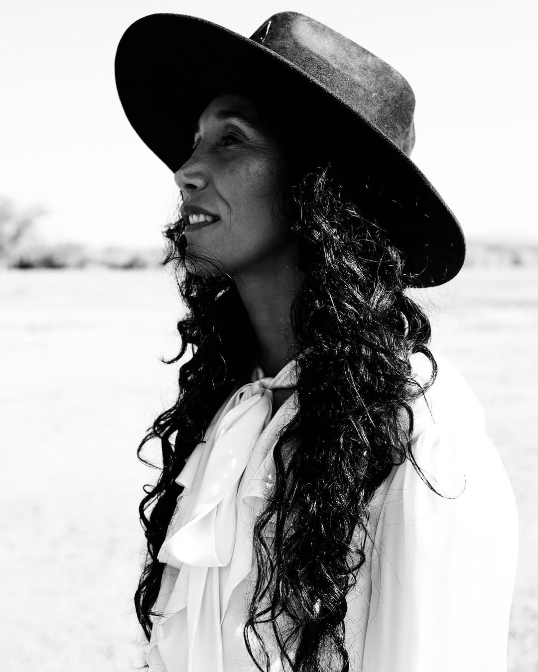 Monochrome portrait of a woman with long curly hair wearing a wide-brimmed hat and white blouse, shot outdoors.