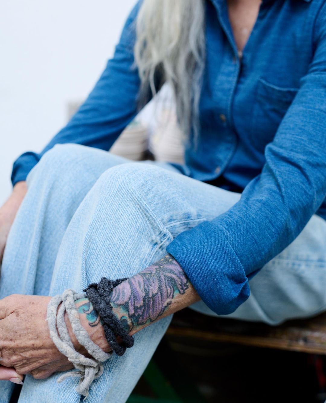 Woman with gray hair and a denim shirt seated with braided bracelets on her wrist, photographed outdoors in soft light.