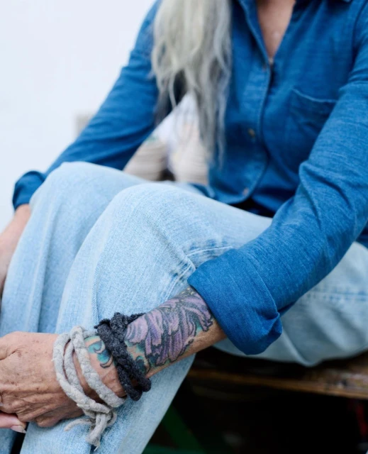 Woman with gray hair and a denim shirt seated with braided bracelets on her wrist, photographed outdoors in soft light.
