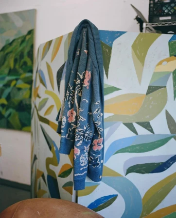 Blue cardigan with a floral pattern hangs against an abstract mural of foliage in a room with an art gallery aesthetic.