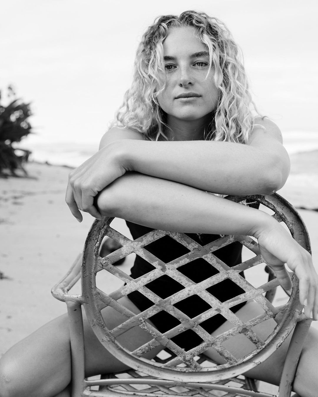 Black and white portrait of a young woman with curly hair in a swimsuit, seated on a chair at the beach with the ocean in the background.