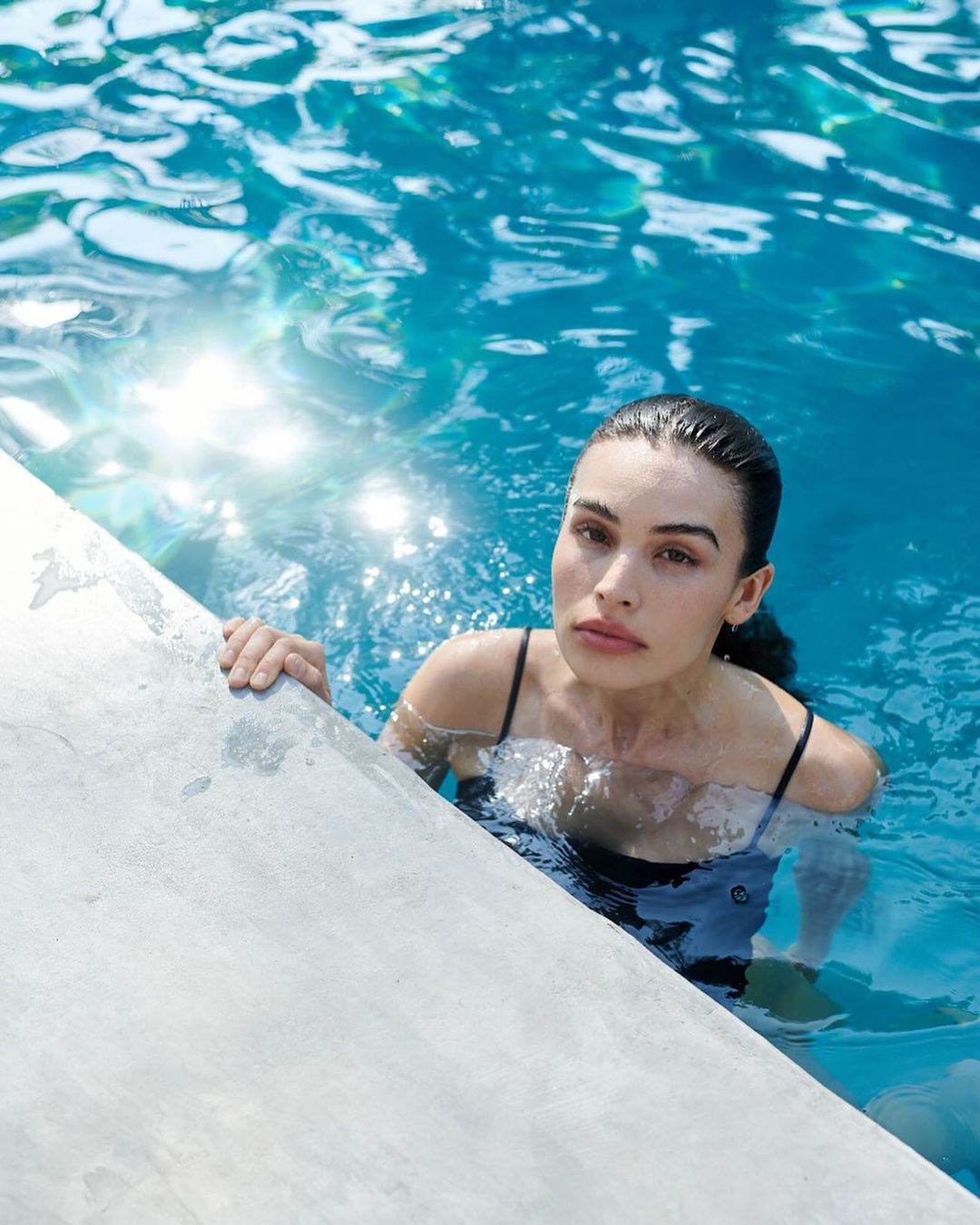 Woman with dark hair and fair skin, partially submerged in a turquoise swimming pool, holds onto the pool's concrete edge, looking directly at the camera.